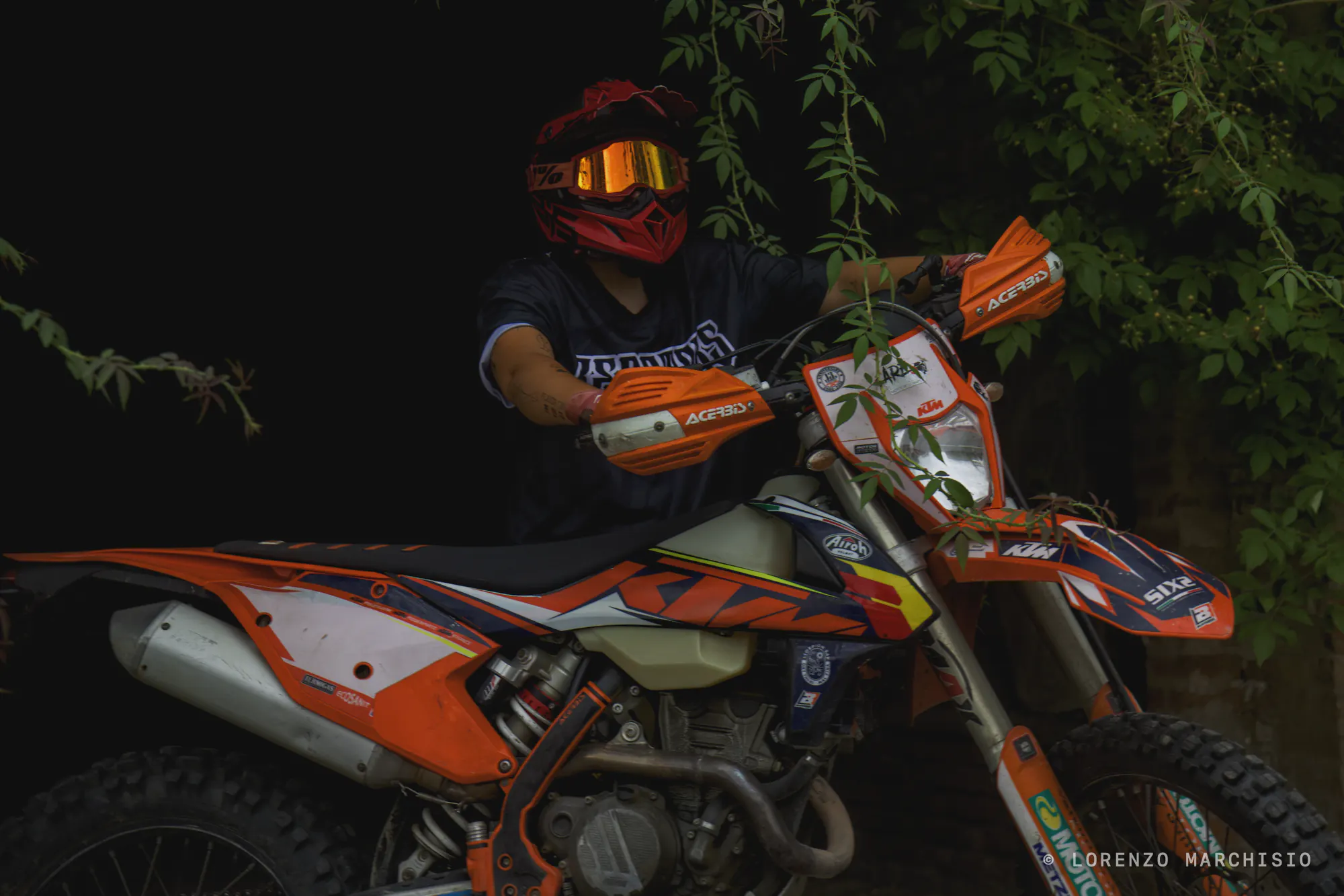 Rider in a red helmet sitting on an orange enduro bike under hanging leaves, lit against the dark